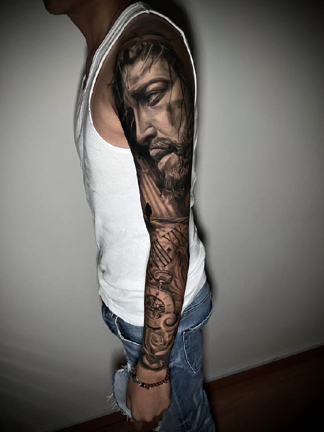 tatuaje realista de rostro de cristo en todo el brazo con rosas y brujula sombras